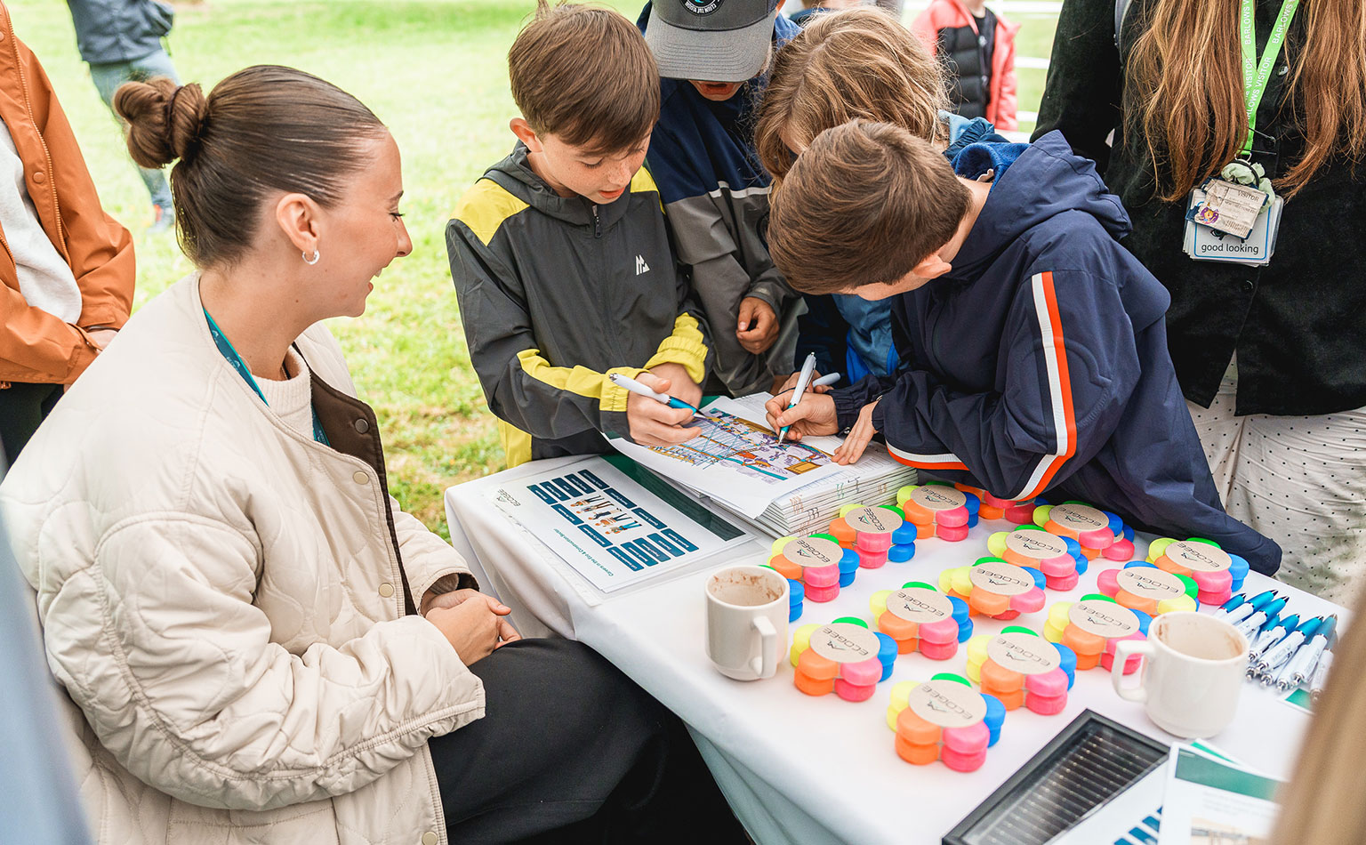 A group of young children crowded around a desk working on activity sheet whilst supervised by an adult. The table is covered with a selection of free, branded items from a Community Threads sponsor.